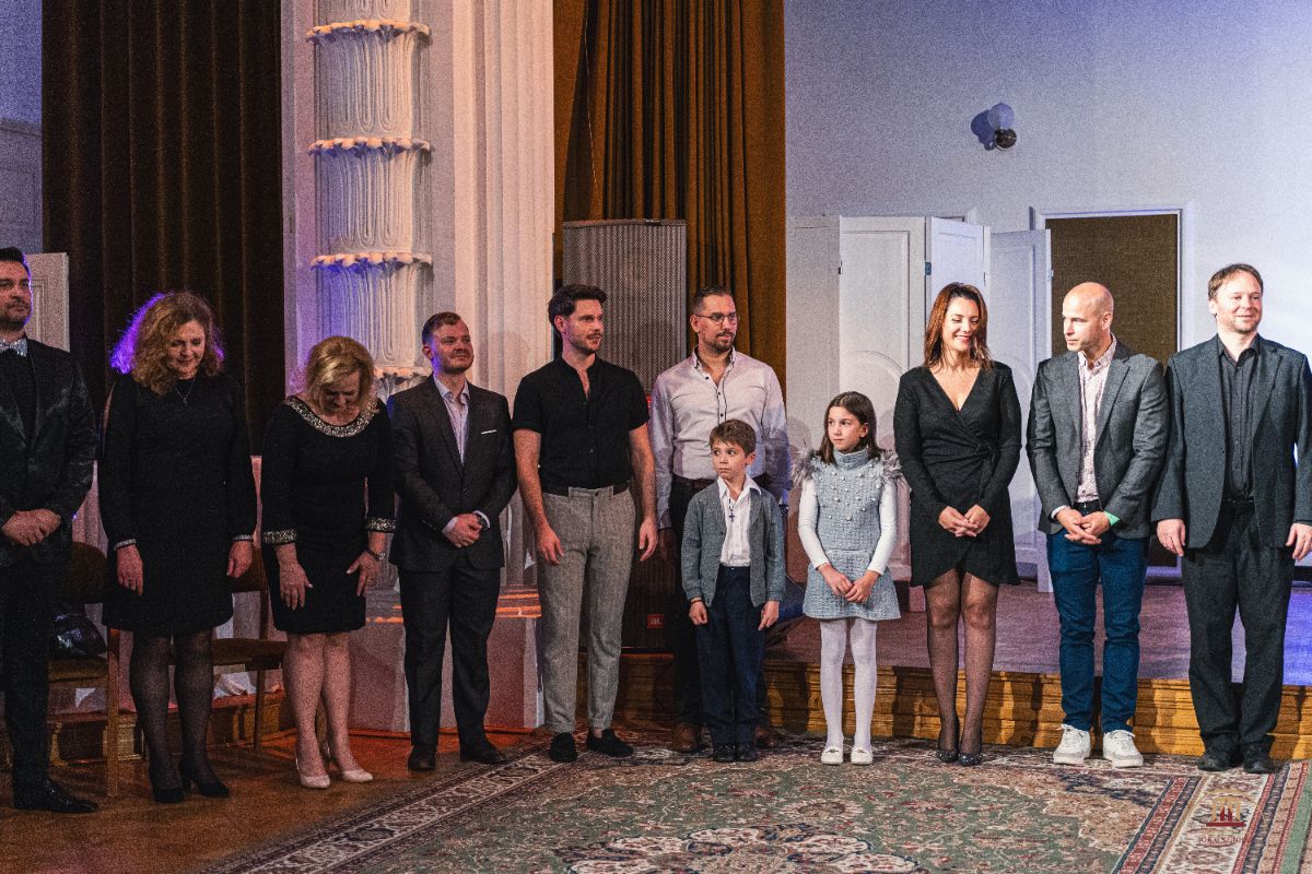 Emberek sora áll egy színpadon elegáns ruhában, köztük két kisgyermek is. A háttérben fehér ajtók és barna függönyök láthatók, a hangulat ünnepélyes.
